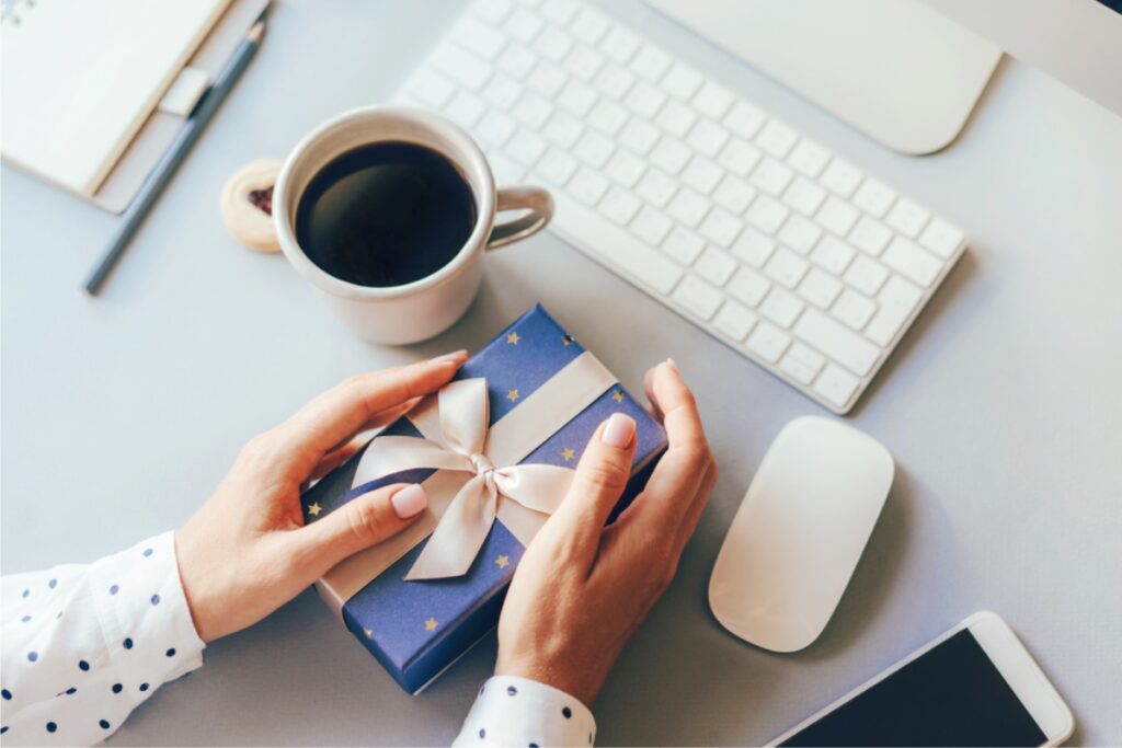 A close-up of a woman's hands presenting a gift box on a desk, with a laptop and coffee cup in the background