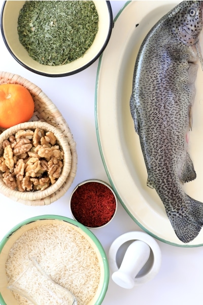 A whole fish on a plate surrounded by bowls containing dried herbs, saffron, rice, walnuts, an orange
