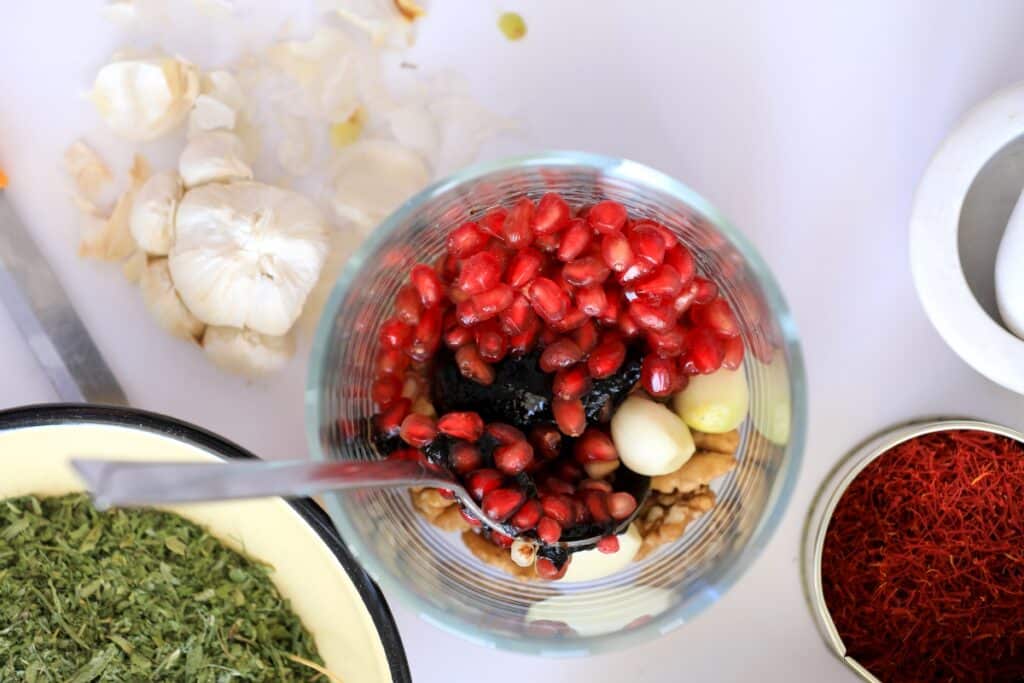 A glass bowl contains pomegranate seeds, walnuts, and garlic cloves. Surrounding it are saffron threads, dried herbs, and garlic on a white surface.