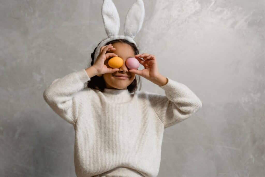 A little girl with bunny ears joyfully holds two colorful eggs in her hands, smiling at the camera.