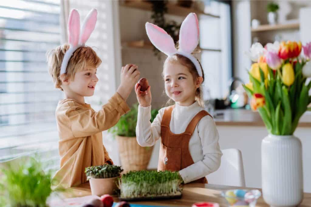 Two children wearing bunny ears, joyfully holding colorful Easter eggs in a festive outdoor setting.