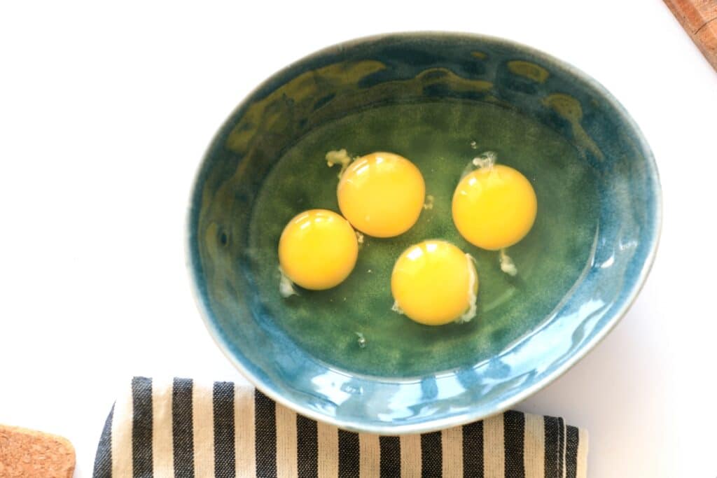 Four eggs in a bowl on a table, accompanied by a knife and a folded towel.