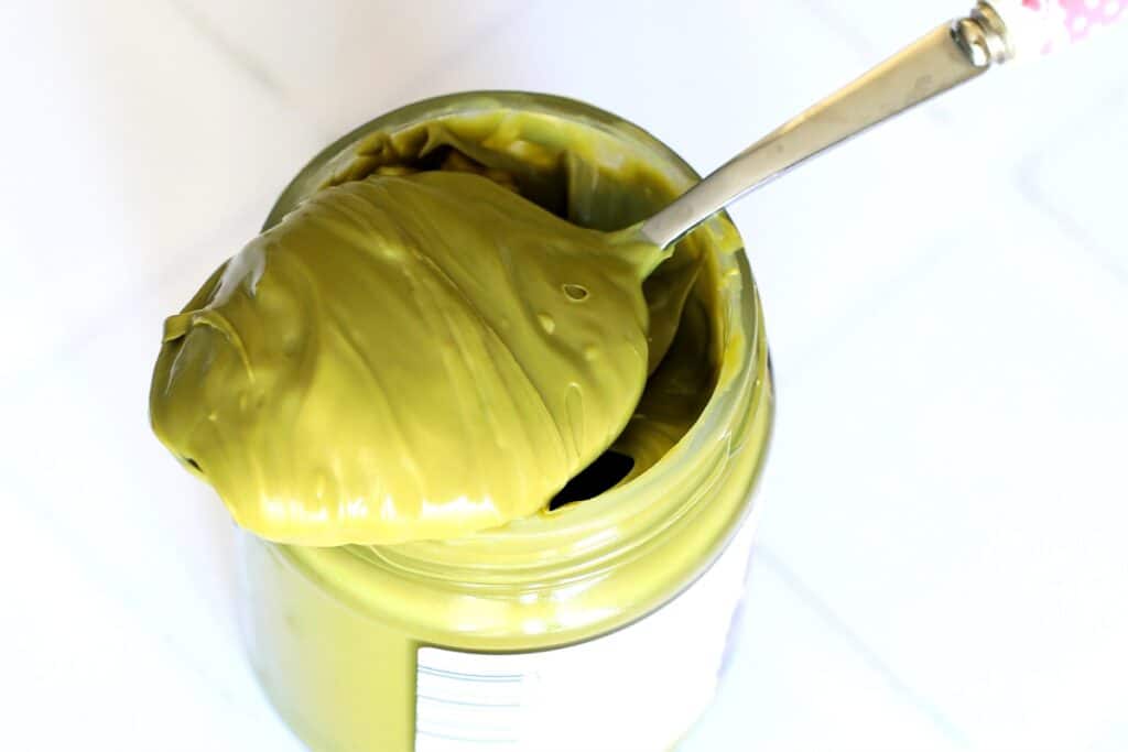 A jar of green paste with a silver spoon resting inside it, placed on a wooden surface.